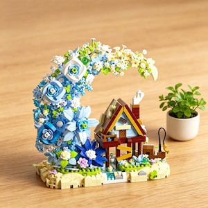 Op de afbeelding: Een miniatuurhuismodel met een bruin dak, blauwe muren en een schoorsteen, omgeven door een grote boog van blauwe en witte bloemen. Rechts staat een kleine potplant. Het model staat op een beige basis.