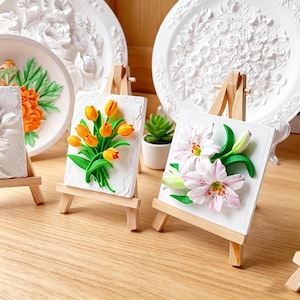 以下が含まれることがあります： 白いレリーフアート作品。花柄のデザイン。木製イーゼルに飾られた2つの小さなアートワークは、オレンジ色のチューリップとピンクのユリのブーケを表示しています。背景には、花柄の装飾的な白いプレートがあります。