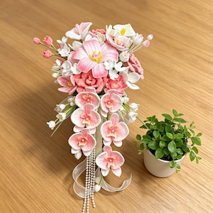 May include: A handcrafted floral arrangement featuring pink and white orchids, tulips, and other flowers. The bouquet is accented with white ribbons and pearl strands. A small potted plant with green leaves is also present.