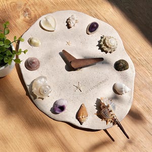 Peut inclure: Une horloge artisanale avec un cadran beige clair de forme irrégulière. L'horloge est ornée de divers coquillages et d'une aiguille de montre en bois foncé. Le texte "PP HANDMADE" est visible. L'horloge est sur une surface en bois.