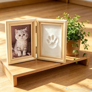 May include: A hinged light wood double frame featuring a photograph of a grey and white kitten on the left side and a white clay paw print impression on the right side, displayed on a matching wooden stand next to a small green potted plant.