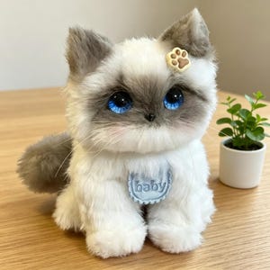 Peut inclure: Un chat en peluche aux yeux bleus vifs, avec une fourrure grise et blanche et un accessoire en forme de patte. Le chat porte un bavoir bleu avec le mot "baby" brodé. Une petite plante en pot est en arrière-plan.