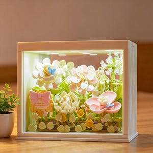 Puede incluir: Una exhibición floral decorativa en un marco rectangular blanco. La exhibición presenta varias flores artificiales en tonos blancos, rosas y amarillos, con follaje verde y pequeñas mariposas. También se incluye un lazo rosa y un pequeño letrero.