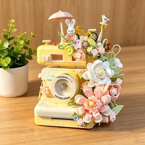 Puede incluir: Una cámara de juguete amarilla decorada con flores, un conejito y una bicicleta en miniatura. La cámara tiene el texto "YAWEL BRICKS" y "YW50mm F1.8" en la parte delantera. Una pequeña sombrilla rosa está encima.