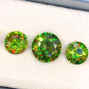 Puede incluir: Tres gemas redondas y facetadas de color verde vibrante. Las gemas muestran destellos internos de color rojo y naranja. Las piedras están sobre un fondo blanco, mostrando su brillo.