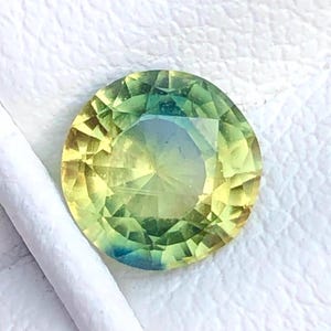 May include: A round, faceted gemstone with a gradient of colors. The gem transitions from yellow to green and blue, with a central, lighter area. The stone is set against a white background, with a white object partially visible.