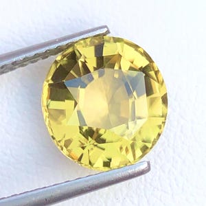Puede incluir: Una gema amarilla facetada, de corte redondo, sujeta con pinzas plateadas. La gema tiene un color brillante y soleado y una superficie brillante y reflectante. Las facetas crean un complejo patrón de luces y sombras.
