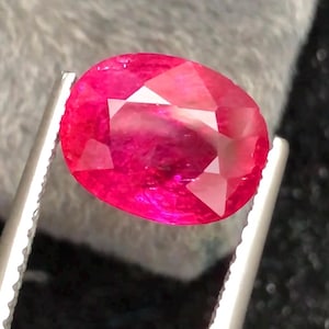 May include: A faceted, oval-cut ruby gemstone, held by silver tweezers. The ruby displays a vibrant, deep red hue with internal reflections. The gemstone is set against a soft, gray background, highlighting its brilliance.