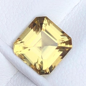 Puede incluir: Una gema facetada, de corte cuadrado, con un tono amarillo cálido. La gema tiene un diseño simétrico con múltiples facetas que reflejan la luz, creando una apariencia brillante. La gema está sobre un fondo blanco.