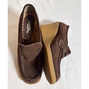 Op de afbeelding: Een paar donkerbruine suède loafers met een sleehak en een lichtbruine rubberen zool. De schoenen hebben witte contraststiksels langs de mocassin-stijl neus en een penny loafer band. De binnenzool is bedrukt met de tekst Paris Blues.