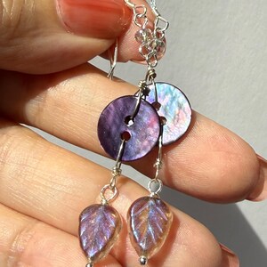 May include: Dangle earrings featuring iridescent purple button-shaped beads and leaf-shaped charms. The earrings have silver-colored hooks and connecting wires. The leaf charms have a translucent, textured appearance with purple and gold hues.