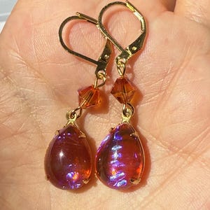 May include: A pair of gold-toned leverback earrings. Each earring features a teardrop-shaped, iridescent, red-purple stone with gold-colored accents. Above the stone hangs a small, orange, faceted crystal. The earrings are displayed in a hand.