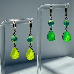 May include: Two pairs of drop earrings featuring faceted lime green teardrop crystals suspended below smaller, round, iridescent blue-green beads. The earrings are attached to bronze-toned lever-back hooks and hang from clear acrylic display stands.