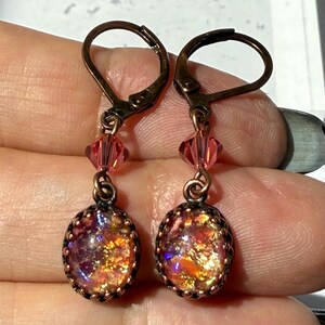 May include: A pair of handcrafted dangle earrings. Each earring features an oval-shaped, iridescent glass cabochon in a bronze-colored setting. The cabochons display a mix of colors, including gold, orange, and purple. The earrings have lever-back closures and are accented with pink crystal beads.
