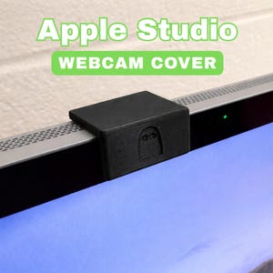 Könnte beinhalten: Eine schwarze Webcam-Abdeckung mit der Aufschrift "Apple Studio" und "WEBCAM COVER" ist an einem Computermonitor befestigt. Die Abdeckung ist rechteckig mit einer strukturierten Oberfläche und einem kleinen Design auf der Vorderseite.