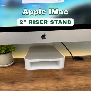 May include: A white 2-inch riser stand for an Apple iMac computer. The stand is rectangular with a hollowed-out center and is positioned beneath the computer's screen. The text "Apple iMac" and "2" RISER STAND" are displayed above the computer.