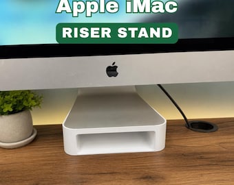 May include: A white Apple iMac riser stand. The stand is rectangular with a hollowed-out section in the middle. The Apple logo is visible on the iMac. The text "Apple iMac RISER STAND" is displayed in green and white.