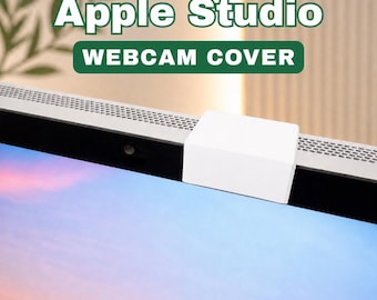 May include: A white webcam cover is placed on top of a computer screen. The text "Apple Studio" and "WEBCAM COVER" are displayed in green and white. The screen displays a gradient of blue and purple.