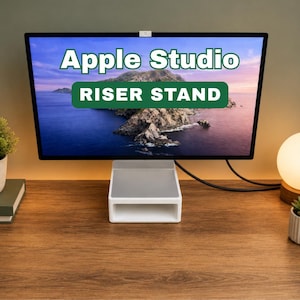 May include: A white and silver riser stand supporting a computer monitor displaying a scenic landscape. The monitor has the text "Apple Studio" and "Riser Stand" in green and white. The stand sits on a wooden desk with a small lamp and plant.