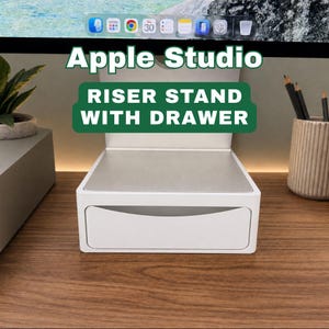 May include: A white Apple Studio riser stand with a drawer. The stand has a silver top and is on a wooden surface. The text "Apple Studio" and "Riser Stand With Drawer" are displayed on the stand.