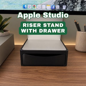 May include: A black Apple Studio riser stand with a drawer. The stand has a silver top and is on a wooden surface. The text "Apple Studio" and "Riser Stand With Drawer" are visible.