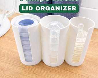 May include: White Philips Avent lid organizer with three compartments. The text "Philips Avent LID ORGANIZER" is displayed on top. Each compartment holds baby bottle parts, including lids, nipples, and bottles. The organizer sits on a wooden surface.