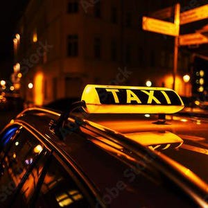 Op de afbeelding: Een close-up van een taxi bij nacht. Op het dak van de auto staat een geel "TAXI" bord met zwarte letters. De achtergrond toont vage lichten en gebouwen.