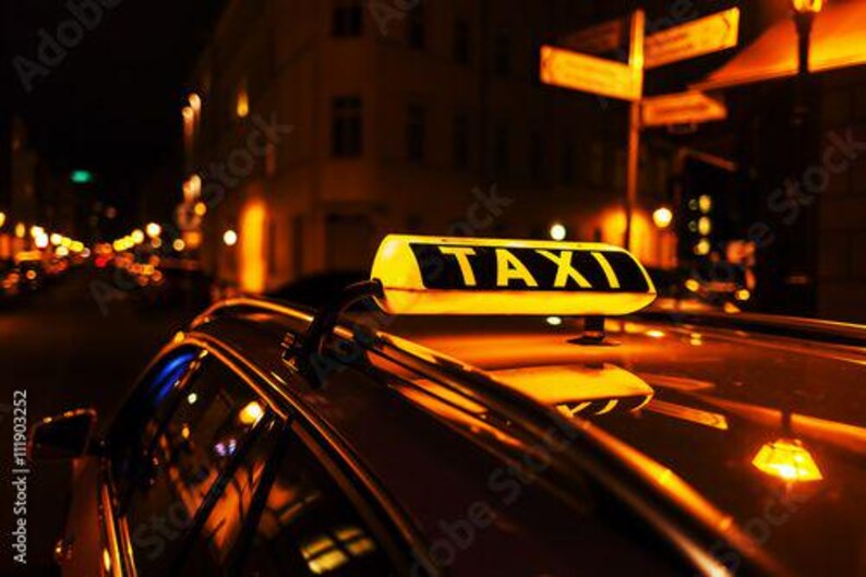 Op de afbeelding: Een close-up van een taxi bij nacht. Op het dak van de auto staat een geel "TAXI" bord met zwarte letters. De achtergrond toont vage lichten en gebouwen.