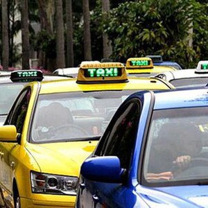Op de afbeelding: Een rij taxi's met verlichte "TAXI" borden op hun daken. De auto's zijn in verschillende kleuren, waaronder geel, zwart en blauw. De foto is overdag genomen, met bomen op de achtergrond.