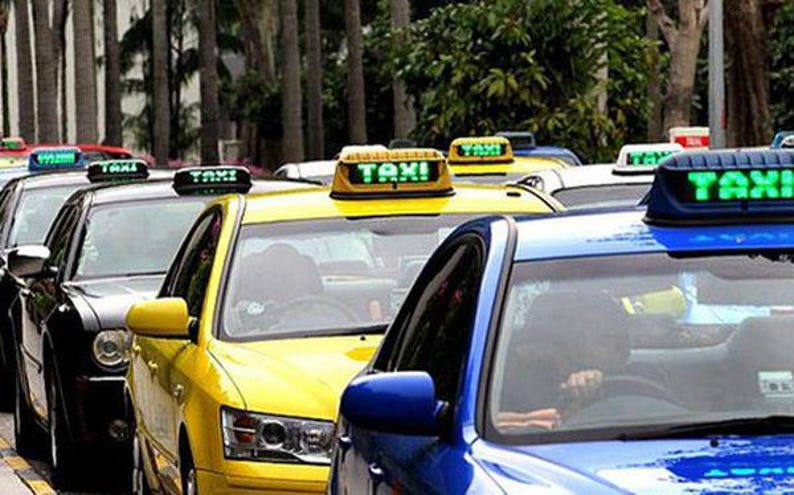 Op de afbeelding: Een rij taxi's met verlichte "TAXI" borden op hun daken. De auto's zijn in verschillende kleuren, waaronder geel, zwart en blauw. De foto is overdag genomen, met bomen op de achtergrond.