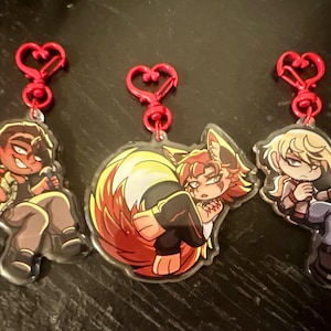 May include: Three acrylic charms with red heart-shaped clasps. Each charm features a cartoon character in various poses. The characters have detailed facial features and are dressed in different outfits. The charms are clear with the character designs printed on them.