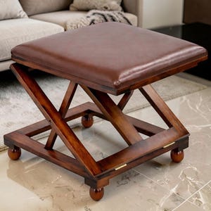 May include: A brown leather-topped ottoman with a dark wood frame. The frame has an X-shaped design and rests on four small, round feet. The ottoman is a square shape and appears to be foldable.
