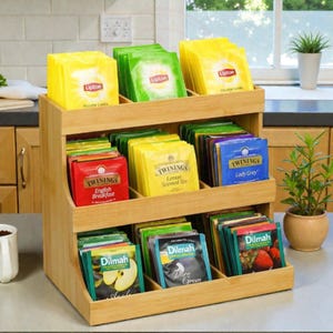 May include: A wooden tea bag organiser with three tiers, filled with tea bags from brands like Lipton, Twinings, and Dilmah. The tea bags are in various colours, including yellow, green, red, and blue. The organiser is made of light-coloured wood and sits on a countertop.
