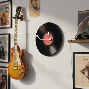 May include: A black vinyl record clock with a red center and white hands. The clock is mounted on a white wall with musical notes. A guitar hangs on the wall to the left, and framed posters are visible.