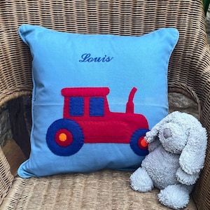 Op de afbeelding: Een blauw kussen met de naam "Louis" geborduurd erop. Een rode vilt tractor met blauwe wielen is op het kussen geplakt. Een grijs knuffeldier zit op het kussen.