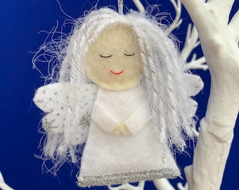 May include: A white felt angel ornament with silver accents. The angel has white yarn hair, closed eyes, and a small red smile. The wings are white with silver glitter. The ornament is hanging against a blue background.