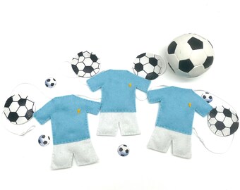 Puede incluir: Tres camisetas de fútbol de fieltro azul con pantalones cortos blancos y una estrella dorada en la parte delantera. Las camisetas están colgadas de una cuerda con balones de fútbol de fieltro blanco unidos.