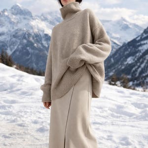 Puede incluir: Un jersey de cuello alto beige y una falda beige a juego. El jersey tiene cuello, puños y dobladillo de canalé. La falda es de largo midi con una abertura delantera. El fondo presenta montañas cubiertas de nieve.