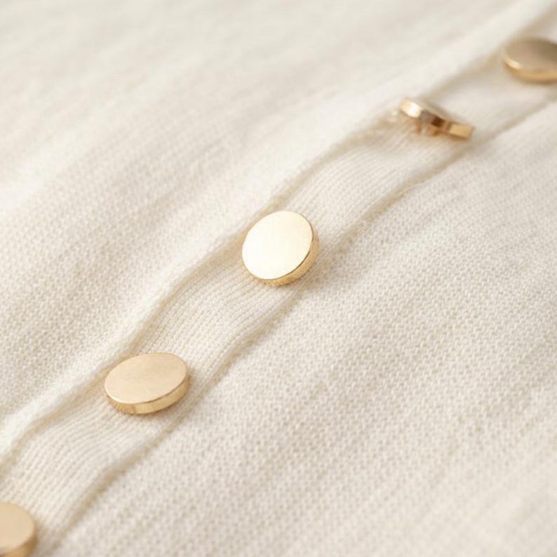 May include: Close-up of a cream-colored knit fabric with a row of gold-colored, round buttons. The buttons are evenly spaced and appear to be attached to a cardigan or sweater.