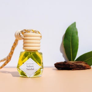 Könnte beinhalten: Kleine, transparente Glasflasche mit Holzverschluss und Seil. Sie enthält eine gelbe Flüssigkeit und trägt das Etikett "Velvet Sandal". Zwei grüne Blätter und ein Stück Rinde liegen daneben. Der Hintergrund ist weiß und pfirsichfarben.