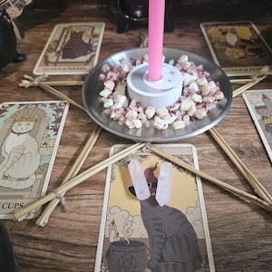 May include: A silver metal candle holder with a central pink taper candle, surrounded by small white and pink stone chips. The holder sits in the center of a star shape formed by five wooden sticks, with tarot cards featuring cat illustrations.
