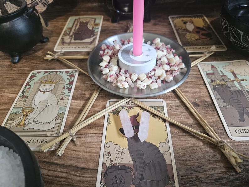 May include: A silver metal candle holder with a central pink taper candle, surrounded by small white and pink stone chips. The holder sits in the center of a star shape formed by five wooden sticks, with tarot cards featuring cat illustrations.