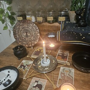 May include: A mystical scene featuring tarot cards with cat illustrations, candles, and a wooden table. Jars of herbs and spices are on a shelf above. A wooden wheel of the year is also present, creating a spiritual atmosphere.