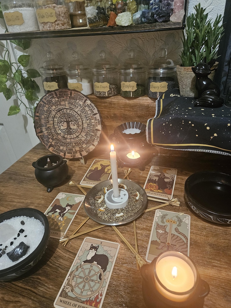 May include: A mystical scene featuring tarot cards with cat illustrations, candles, and a wooden table. Jars of herbs and spices are on a shelf above. A wooden wheel of the year is also present, creating a spiritual atmosphere.