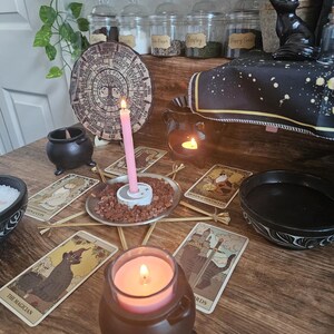 May include: A wooden table surface featuring a circular calendar wheel, tarot cards, a pink candle in a white holder surrounded by red gravel, a brown cauldron with a pink candle, a black bowl, and a black oil burner with a lit tea light candle.
