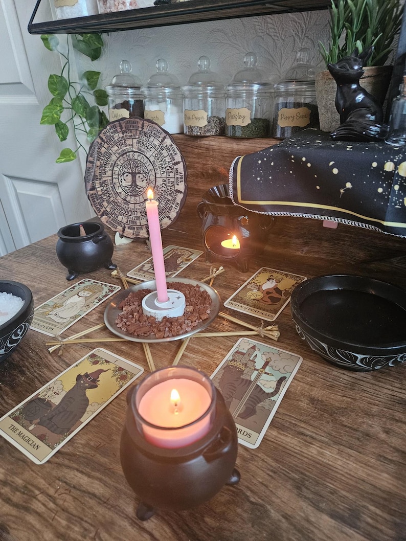 May include: A wooden table surface featuring a circular calendar wheel, tarot cards, a pink candle in a white holder surrounded by red gravel, a brown cauldron with a pink candle, a black bowl, and a black oil burner with a lit tea light candle.