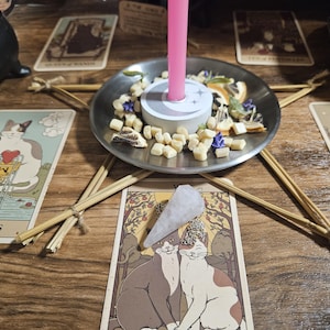 May include: A pink candle stands in a silver metal dish filled with dried herbs and small white cubes. A clear crystal pendulum rests on a tarot card featuring two cats. A star-shaped frame made of thin wooden sticks surrounds the central dish.