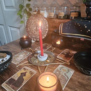 May include: A ritual setup featuring a red candle in a white holder centered on a star-shaped twig frame, surrounded by tarot cards including The Magician and Six of Pentacles, small black cauldrons with candles, and a circular wooden calendar.
