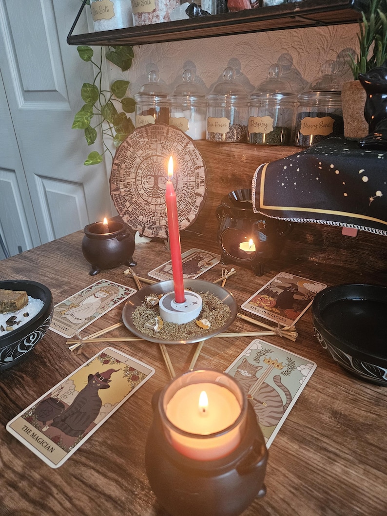 May include: A ritual setup featuring a red candle in a white holder centered on a star-shaped twig frame, surrounded by tarot cards including The Magician and Six of Pentacles, small black cauldrons with candles, and a circular wooden calendar.