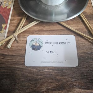 May include: A white rectangular card featuring a circular logo with a cat in a cauldron, the text "One Spoon", "With love and gratitude", and "onespoonwitch.com". The card is decorated with small black stars and moon phase symbols.